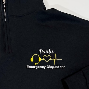 May include: Black quarter-zip sweatshirt with a yellow embroidered design. The design includes a headset, a heart, and a heartbeat line. The text "Paula Emergency Dispatcher" is embroidered below the design.