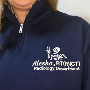 May include: Navy blue quarter-zip sweatshirt with white embroidered skeleton graphic and text. The text reads "Alesha, RT(R)(CT) Radiology Department". The sweatshirt has a silver zipper and a ribbed collar.