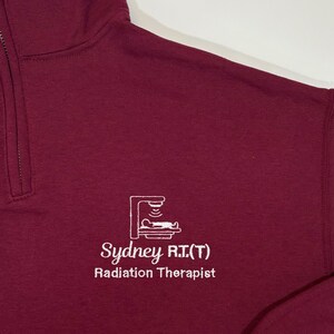 May include: Burgundy colored long-sleeve sweatshirt with a white embroidered logo that says "Sydney R.T.(T) Radiation Therapist".