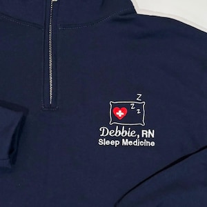 May include: Navy blue quarter-zip sweatshirt with a silver zipper. The sweatshirt features an embroidered design of a red heart with a white cross, two Z's, and the text "Debbie, RN Sleep Medicine" in white.