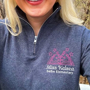 K&ouml;nnte beinhalten: Ein dunkelgrauer Rei&szlig;verschluss-Pullover mit einem rosa gestickten Design, das ein Buch mit Sternen und den Text "Miss Kelsea Selbe Elementary" zeigt.