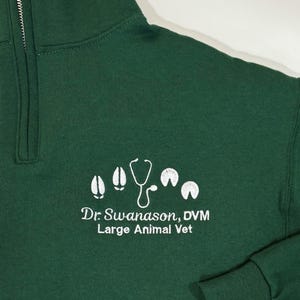 May include: A dark green, quarter-zip sweatshirt with a white embroidered design. The design includes a stethoscope, four hoof prints, and the text "Dr. Swanson, DVM Large Animal Vet".