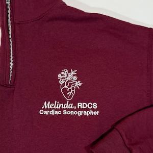 May include: Burgundy colored sweatshirt with a white embroidered heart with flowers and the text "Melinda, RDCS Cardiac Sonographer".
