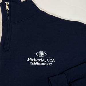 May include: A dark blue quarter-zip sweatshirt with a white embroidered eye logo and the text "Michaela, COA Ophthalmology".