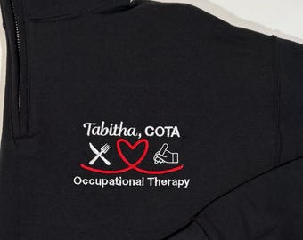 May include: Black quarter-zip sweatshirt with white embroidery that reads "Tabitha, COTA" and "Occupational Therapy". The embroidery also includes a red heart with a fork and knife on the left and a hand with a pencil on the right.