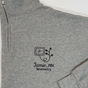 May include: Gray quarter-zip sweatshirt with a black embroidered design and text. The design includes a heart rate monitor symbol and the text "Jamie, RN Telemetry". The sweatshirt has a high collar and long sleeves.