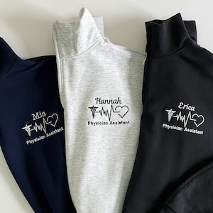 May include: Three zippered sweatshirts in navy blue, gray, and black with a white embroidered design of a caduceus, a heart, and a heartbeat line. Each sweatshirt has a different name embroidered below the design: Mia, Hannah, and Erica. The text below the design reads "Physician Assistant".