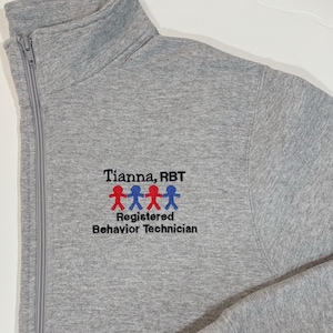 May include: Gray zippered sweatshirt with a black embroidered logo that says "Tianna, RBT" and "Registered Behavior Technician" with a row of four red and blue stick figures.