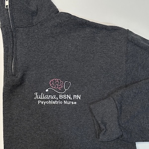 May include: A dark gray 1/4 zip sweatshirt with a white embroidered brain and stethoscope design. The text "Juliana, BSN, RN Psychiatric Nurse" is also embroidered in white.