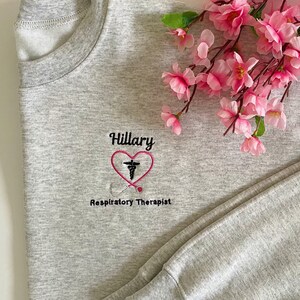 May include: Gray sweatshirt with a pink heart and a stethoscope design embroidered on the front. The text "Hillary Respiratory Therapist" is embroidered below the heart.