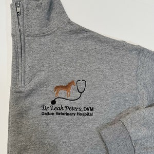 May include: Gray zippered sweatshirt with a brown horse and stethoscope logo embroidered on the front. The text "Dr. Leah Peters, DVM Dalton Veterinary Hospital" is also embroidered below the logo.