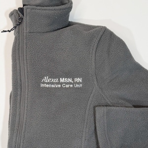 May include: A gray fleece jacket with a full zipper and a high collar. The jacket has the embroidered text "Alexa MSN, RN Intensive Care Unit" in white. The jacket is folded on a white surface.