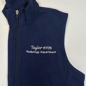 May include: A navy blue fleece vest with a zipper closure. The vest has the text "Taylor RT(R) Radiology Department" embroidered on the front in white.