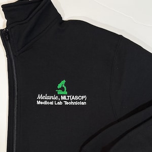 May include: Black zippered jacket with a green microscope logo and white text that reads "Melanie, MLT(ASCP) Medical Lab Technician".