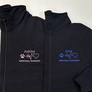 May include: Two black zip-up jackets with embroidered designs. One jacket features the name "Ashley" above a paw print, heartbeat line, and heart, with "Veterinary Assistant" below. The other jacket has "Alex" with a similar design in blue.