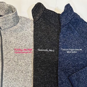 May include: Three fleece zip-up jackets in gray, black, and navy blue. Each jacket has a name and title embroidered on the chest. The gray jacket has pink embroidery, the black jacket has white embroidery, and the navy jacket has white embroidery.