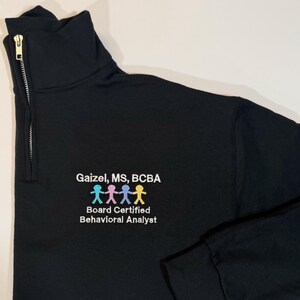 May include: Black quarter-zip sweatshirt with a gold zipper. The sweatshirt features the text "Gaizel, MS, BCBA" and a colorful graphic of stick figures. Below the text is "Board Certified Behavioral Analyst".