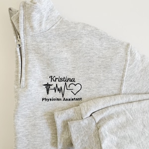 May include: Gray zippered sweatshirt with a black embroidered design featuring a medical symbol, a heart, and a heartbeat line. The text "Kristina Physician Assistant" is also embroidered on the sweatshirt.