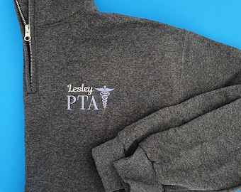 Puede incluir: Una sudadera gris oscuro con cremallera de cuarto de cuello con el texto bordado "Lesley PTA" y un símbolo médico blanco. La sudadera tiene una cremallera plateada y mangas dobladas. El fondo es azul liso.