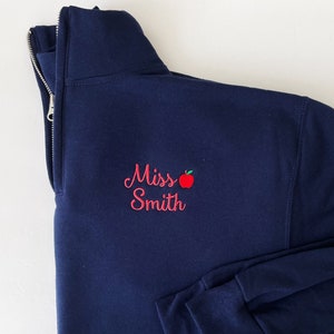 May include: A navy blue sweatshirt with a zipper and a red embroidered apple and the text "Miss Smith" on the front.