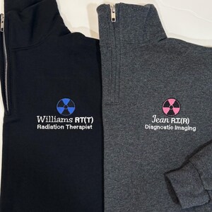 May include: Two quarter-zip sweatshirts, one black and one gray. The black sweatshirt has blue text and a blue radiation symbol that reads "Williams RT(T) Radiation Therapist." The gray sweatshirt has pink text and a pink radiation symbol that reads "Jean R.T.(R) Diagnostic Imaging."