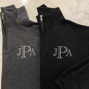May include: Two quarter-zip pullovers, one charcoal gray and one black, are displayed. Each features a white embroidered "JPA" monogram on the chest. The pullovers have a mock neck and a silver zipper.