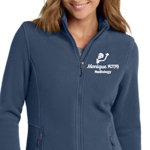 May include: A navy blue fleece jacket with a full zipper and two side pockets. The jacket features the text "Monique RT(R) Radiology" in white on the upper left chest. The jacket is a comfortable and warm outerwear option.