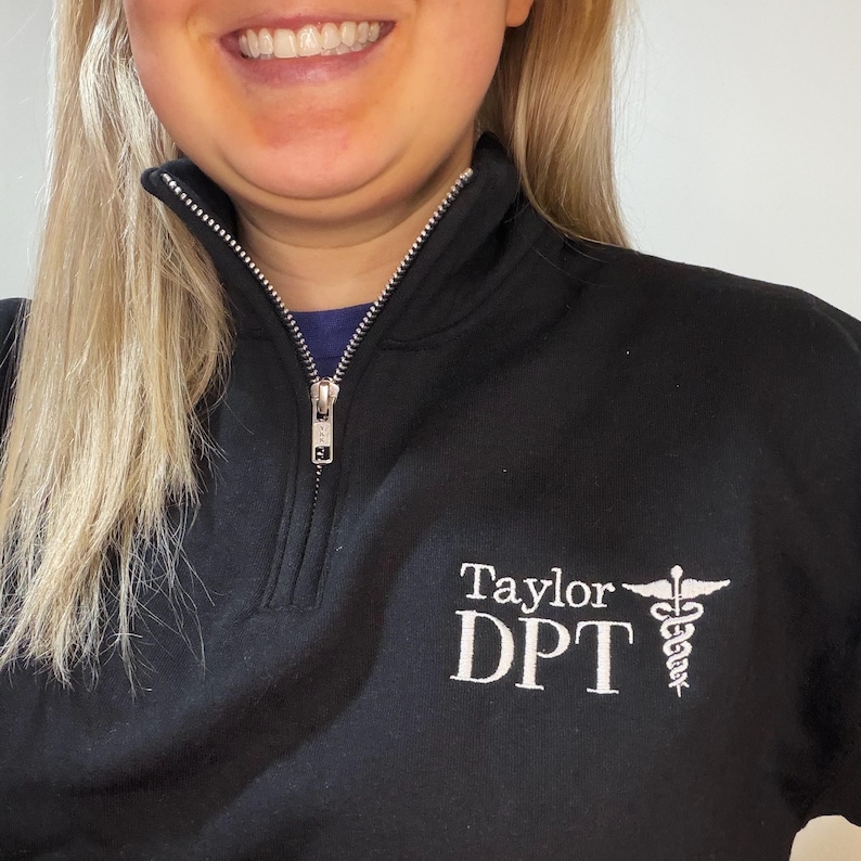 Puede incluir: Sudadera negra con cremallera de cuarto de cremallera y cremallera plateada. Texto bordado en blanco "Taylor DPT" y un s&iacute;mbolo m&eacute;dico. La sudadera tiene un cuello alto y est&aacute; hecha de un material suave.