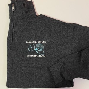 May include: Dark gray quarter-zip sweatshirt with the text "Madison, BSN, RN" above a light blue embroidered design of a brain, stethoscope, and the words "Psychiatric Nurse".