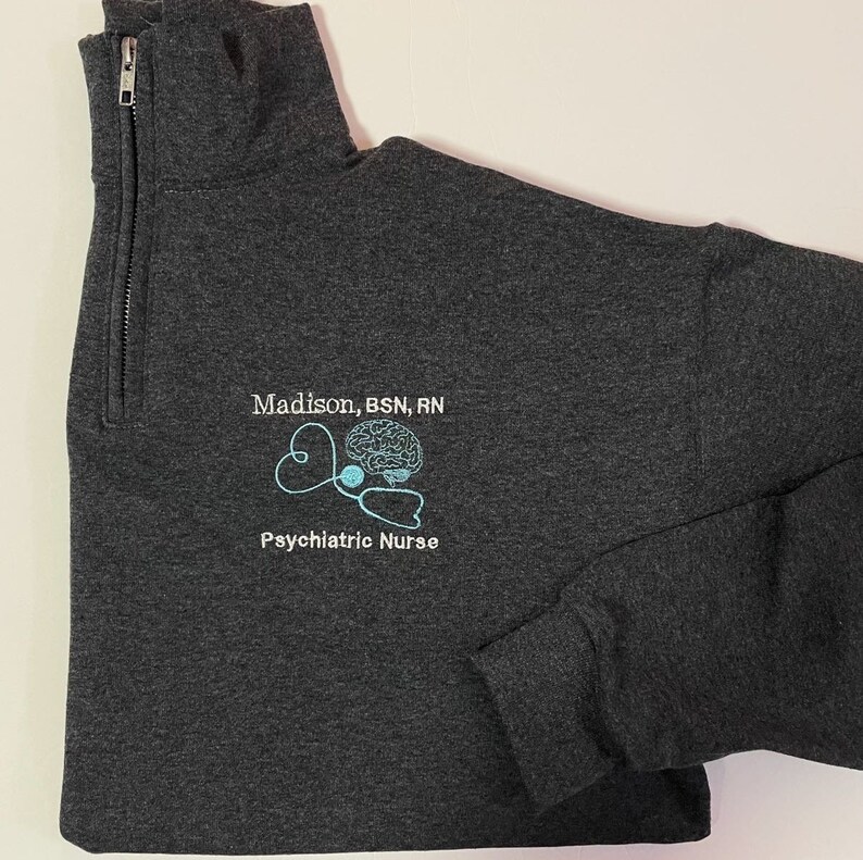 May include: Dark gray quarter-zip sweatshirt with the text "Madison, BSN, RN" above a light blue embroidered design of a brain, stethoscope, and the words "Psychiatric Nurse".