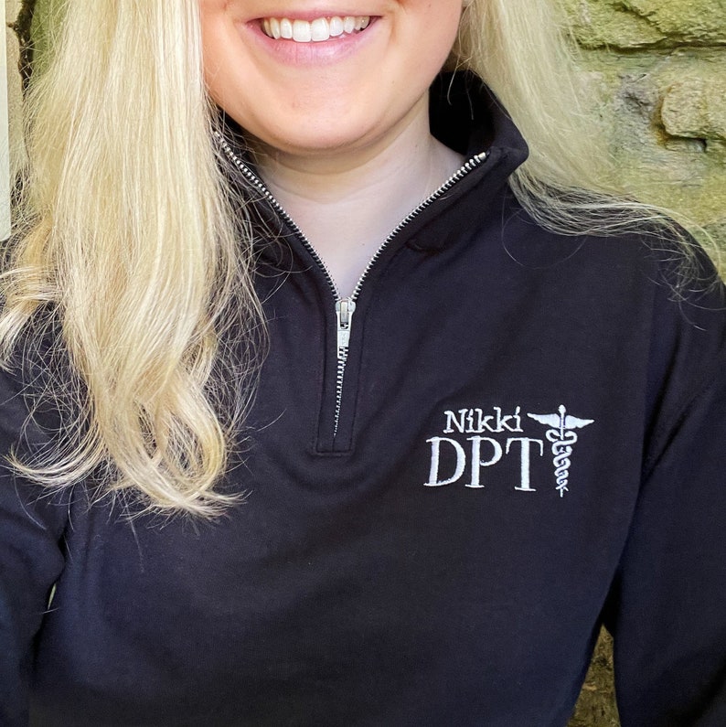 Puede incluir: Sudadera negra con cremallera de cuarto de cuello con un logotipo blanco bordado que dice "Nikki DPT" y un s&iacute;mbolo m&eacute;dico. La sudadera tiene una cremallera plateada y un cuello alto.