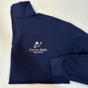 Pode incluir: Camisola azul marinho com fecho de correr de um quarto de gola, com um desenho de esqueleto branco bordado e o texto "Juwan R.I.(R) Rad Tech". A camisola tem gola alta e mangas compridas.