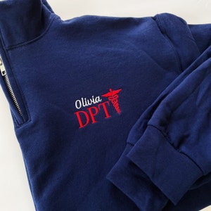 May include: A navy blue quarter-zip sweatshirt featuring embroidered text that reads Olivia DPT in white and red lettering, accompanied by a red caduceus symbol. The garment has long sleeves with ribbed cuffs and a silver-toned metal zipper.