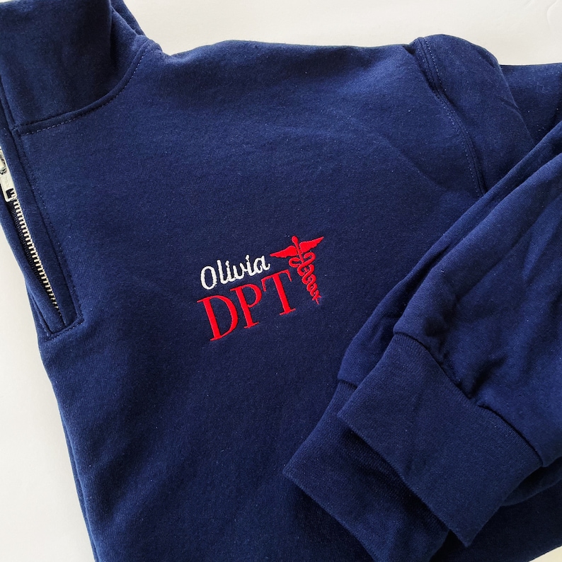 May include: A navy blue quarter-zip sweatshirt featuring embroidered text that reads Olivia DPT in white and red lettering, accompanied by a red caduceus symbol. The garment has long sleeves with ribbed cuffs and a silver-toned metal zipper.