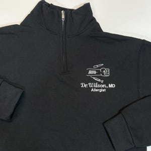 Pode incluir: Camisola preta com fecho de correr de um quarto com um gr&aacute;fico branco de um punho, uma seringa e s&iacute;mbolos m&eacute;dicos. O texto "Dr. Wilson, MD Allergist" est&aacute; impresso abaixo.