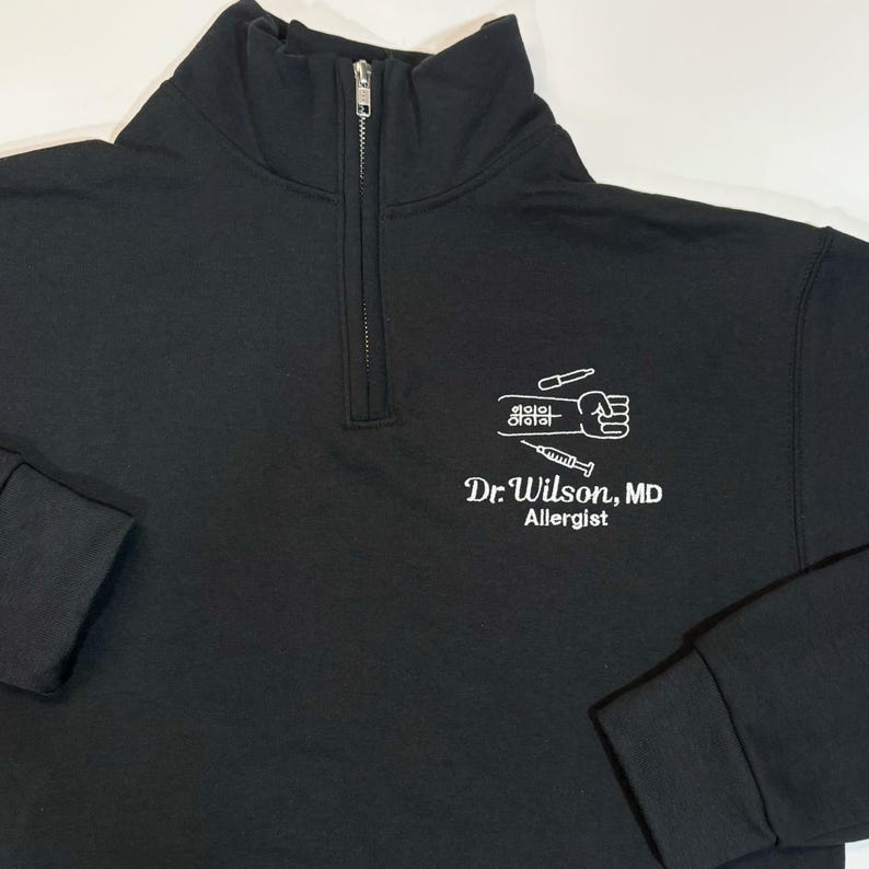 Pode incluir: Camisola preta com fecho de correr de um quarto com um gr&aacute;fico branco de um punho, uma seringa e s&iacute;mbolos m&eacute;dicos. O texto "Dr. Wilson, MD Allergist" est&aacute; impresso abaixo.