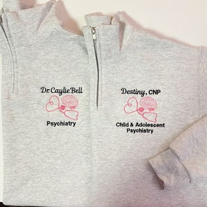 May include: Light gray quarter-zip sweatshirt with embroidered pink brain and stethoscope design. The sweatshirt features the text "Dr. Caylie Bell Psychiatry" and "Destiny, CNP Child & Adolescent Psychiatry".