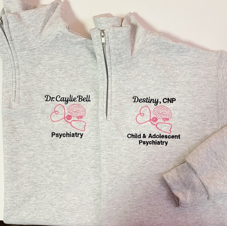 May include: Light gray quarter-zip sweatshirt with embroidered pink brain and stethoscope design. The sweatshirt features the text "Dr. Caylie Bell Psychiatry" and "Destiny, CNP Child & Adolescent Psychiatry".