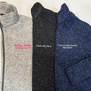 K&ouml;nnte beinhalten: Drei Fleece-Jacken mit Rei&szlig;verschluss in Grau, Schwarz und Marineblau. Jede Jacke hat einen hohen Kragen und ist mit einem Namen und einem beruflichen Titel in wei&szlig;em oder rosa Faden bestickt. Die Jacken bestehen aus einem weichen, strukturierten Material.