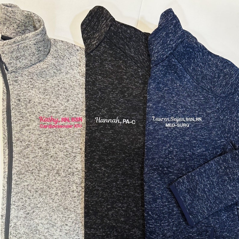 K&ouml;nnte beinhalten: Drei Fleece-Jacken mit Rei&szlig;verschluss in Grau, Schwarz und Marineblau. Jede Jacke hat einen hohen Kragen und ist mit einem Namen und einem beruflichen Titel in wei&szlig;em oder rosa Faden bestickt. Die Jacken bestehen aus einem weichen, strukturierten Material.