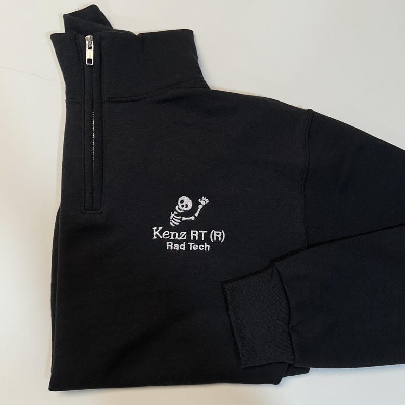 Pode incluir: Camisola preta com fecho de correr a um quarto, gola alta e fecho de correr prateado. A camisola apresenta texto bordado branco e um gr&aacute;fico de esqueleto que diz "Kenz RT (R) Rad Tech". As mangas s&atilde;o compridas e os punhos s&atilde;o canelados.