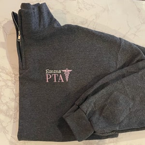 May include: A dark gray quarter-zip sweatshirt with a gold zipper. The left chest features the embroidered text "Emma PTA" in pink, along with a pink caduceus symbol. The sweatshirt has long sleeves and a folded cuff.