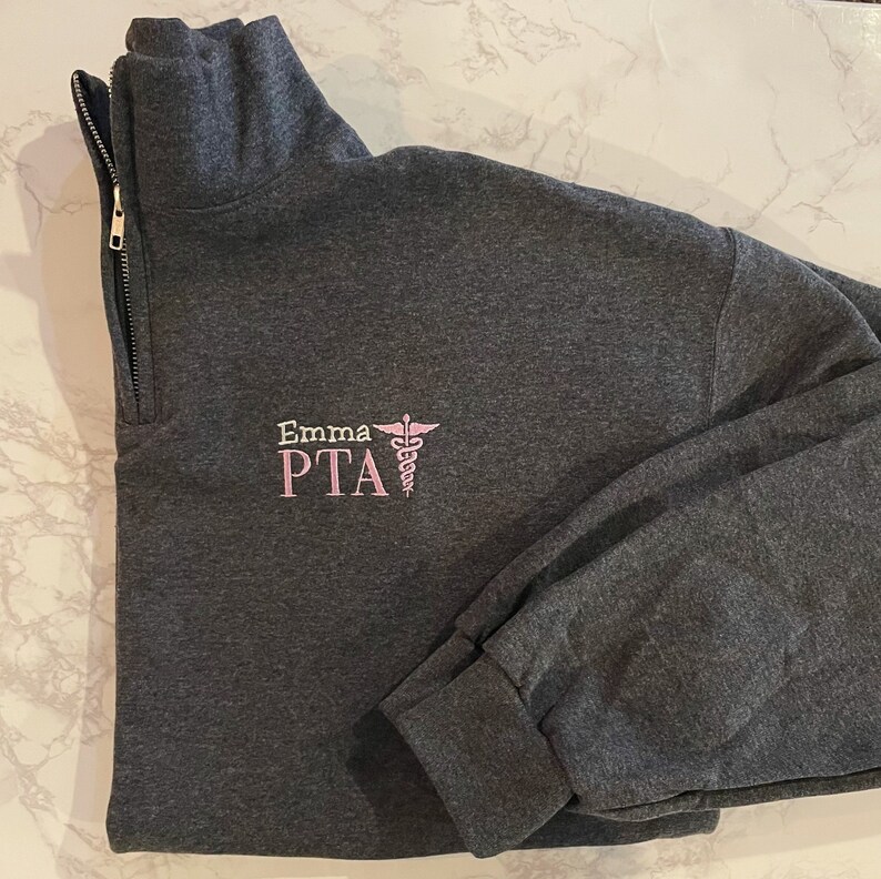 May include: A dark gray quarter-zip sweatshirt with a gold zipper. The left chest features the embroidered text "Emma PTA" in pink, along with a pink caduceus symbol. The sweatshirt has long sleeves and a folded cuff.