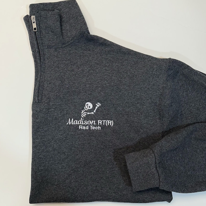 Pode incluir: Camisola cinza escuro com z&iacute;per de um quarto de gola, com um gr&aacute;fico de esqueleto bordado em branco e o texto "Madison RT(R) Rad Tech". A camisola tem uma gola alta e um z&iacute;per prateado.
