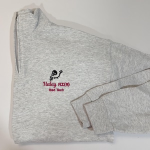 Pode incluir: Um moletom cinza claro com z&iacute;per de um quarto, com um gr&aacute;fico de esqueleto preto e texto rosa que diz "Haley RI(R) Rad Tech". O moletom tem gola alta e mangas compridas.