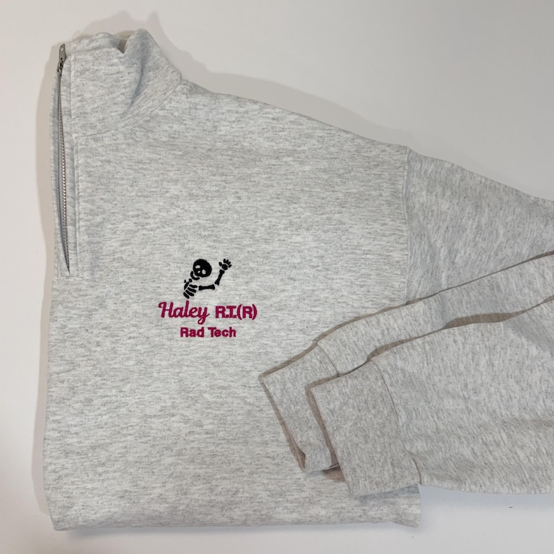 Pode incluir: Um moletom cinza claro com z&iacute;per de um quarto, com um gr&aacute;fico de esqueleto preto e texto rosa que diz "Haley RI(R) Rad Tech". O moletom tem gola alta e mangas compridas.