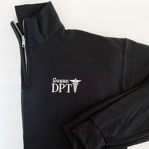 Puede incluir: Sudadera negra con cremallera de cuarto de cuello, texto bordado en blanco y un s&iacute;mbolo m&eacute;dico. El texto dice "Susan DPT". La sudadera tiene un cuello alto y una cremallera plateada.
