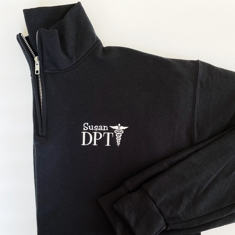 Puede incluir: Sudadera negra con cremallera de cuarto de cuello, texto bordado en blanco y un s&iacute;mbolo m&eacute;dico. El texto dice "Susan DPT". La sudadera tiene un cuello alto y una cremallera plateada.
