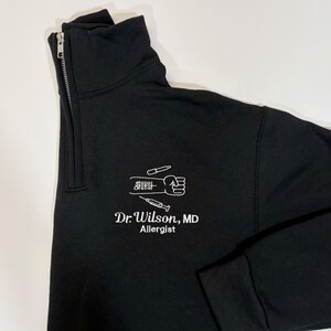 Pode incluir: Camisola preta com fecho de correr a meio do pesco&ccedil;o com um gr&aacute;fico branco de um punho, uma seringa e s&iacute;mbolos m&eacute;dicos. O texto "Dr. Wilson, MD Allergist" est&aacute; abaixo. A camisola tem uma gola alta.