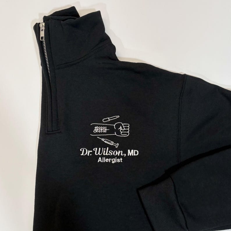 Pode incluir: Camisola preta com fecho de correr a meio do pesco&ccedil;o com um gr&aacute;fico branco de um punho, uma seringa e s&iacute;mbolos m&eacute;dicos. O texto "Dr. Wilson, MD Allergist" est&aacute; abaixo. A camisola tem uma gola alta.